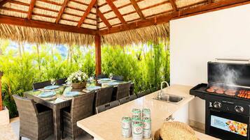 An outdoor dining area with a grill, wicker furniture, and a thatched roof.