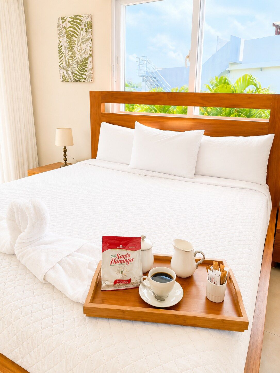 Una cama bien tendida con ropa blanca, una bandeja con cafetera, taza y un paquete de café Santo Domingo, y una ventana con vista a follaje y un edificio.