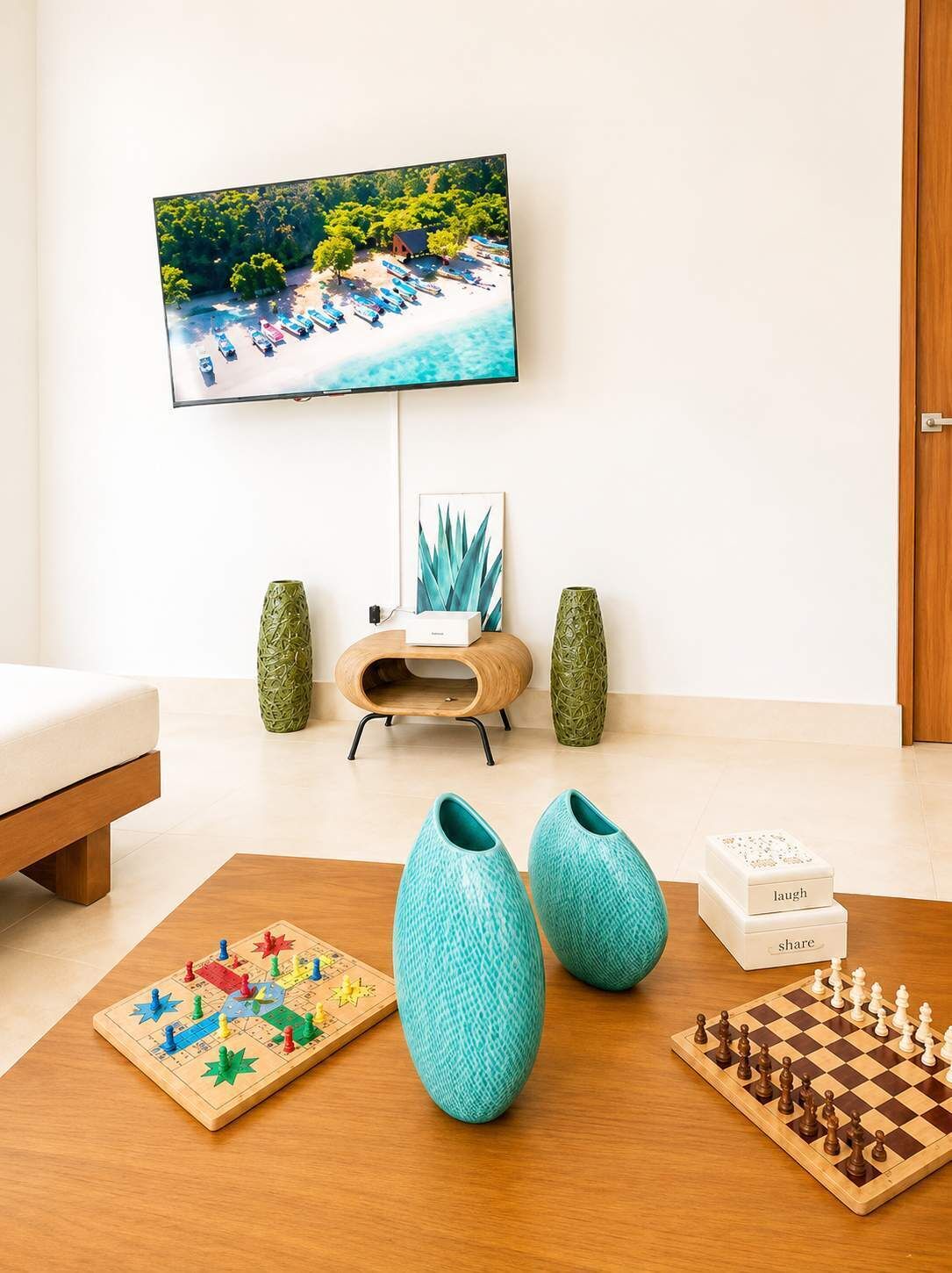 A living room with a television mounted on the wall displaying a beach scene, a chessboard, and two vases on a coffee table.