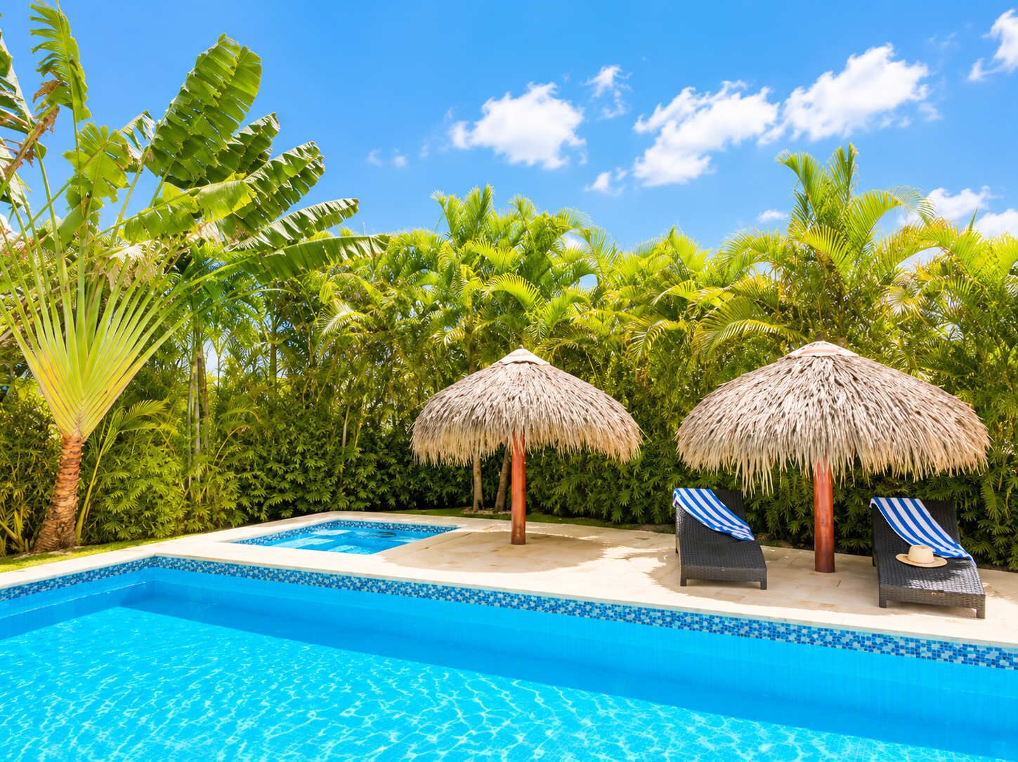 A swimming pool with two lounge chairs and thatched umbrellas, surrounded by lush tropical vegetation.