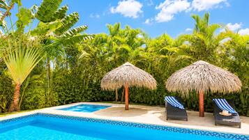 A swimming pool with two lounge chairs and thatched umbrellas, surrounded by lush tropical vegetation.