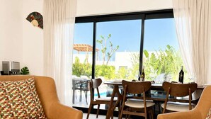 Ein modernes Wohnzimmer mit Schachbrett auf einem Couchtisch, Ledersessel, Essbereich mit Blick ins Freie und einem großen Fenster mit Vorhängen.