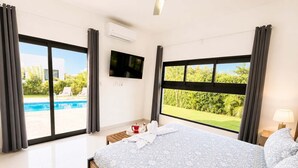 A hotel room with a bed, a TV, a window with a view of a swimming pool and greenery, and a ceiling fan. - 3BD Villa in SOV | Private Pool & Peaceful Retreat (Sosúa)