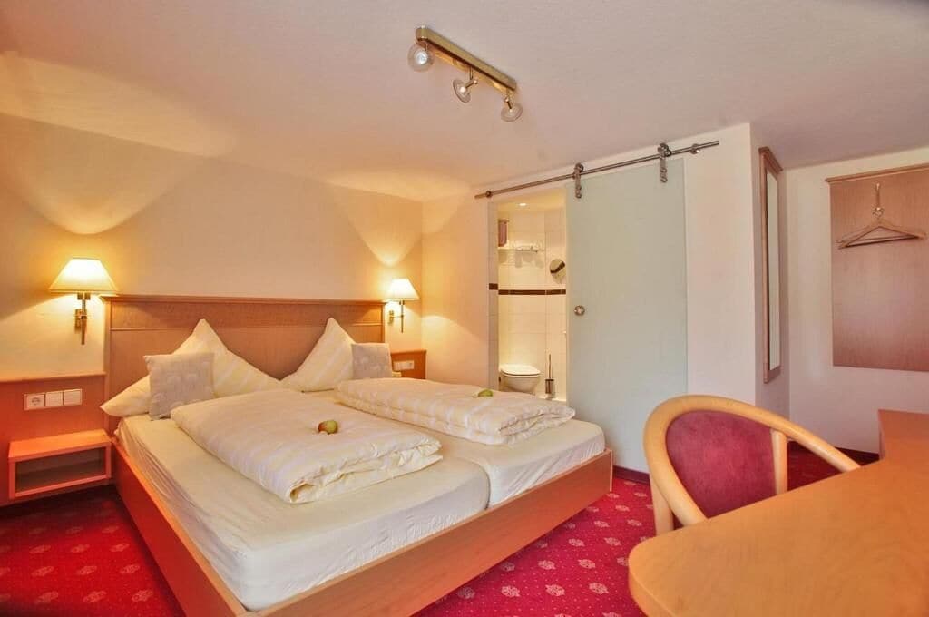 A hotel room with a bed, bedside lamps, a chair, a table, and a wardrobe.