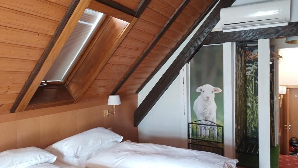 A bedroom with a sloped ceiling, a bed with white linens, a wall-mounted lamp, and a door with a metal gate.