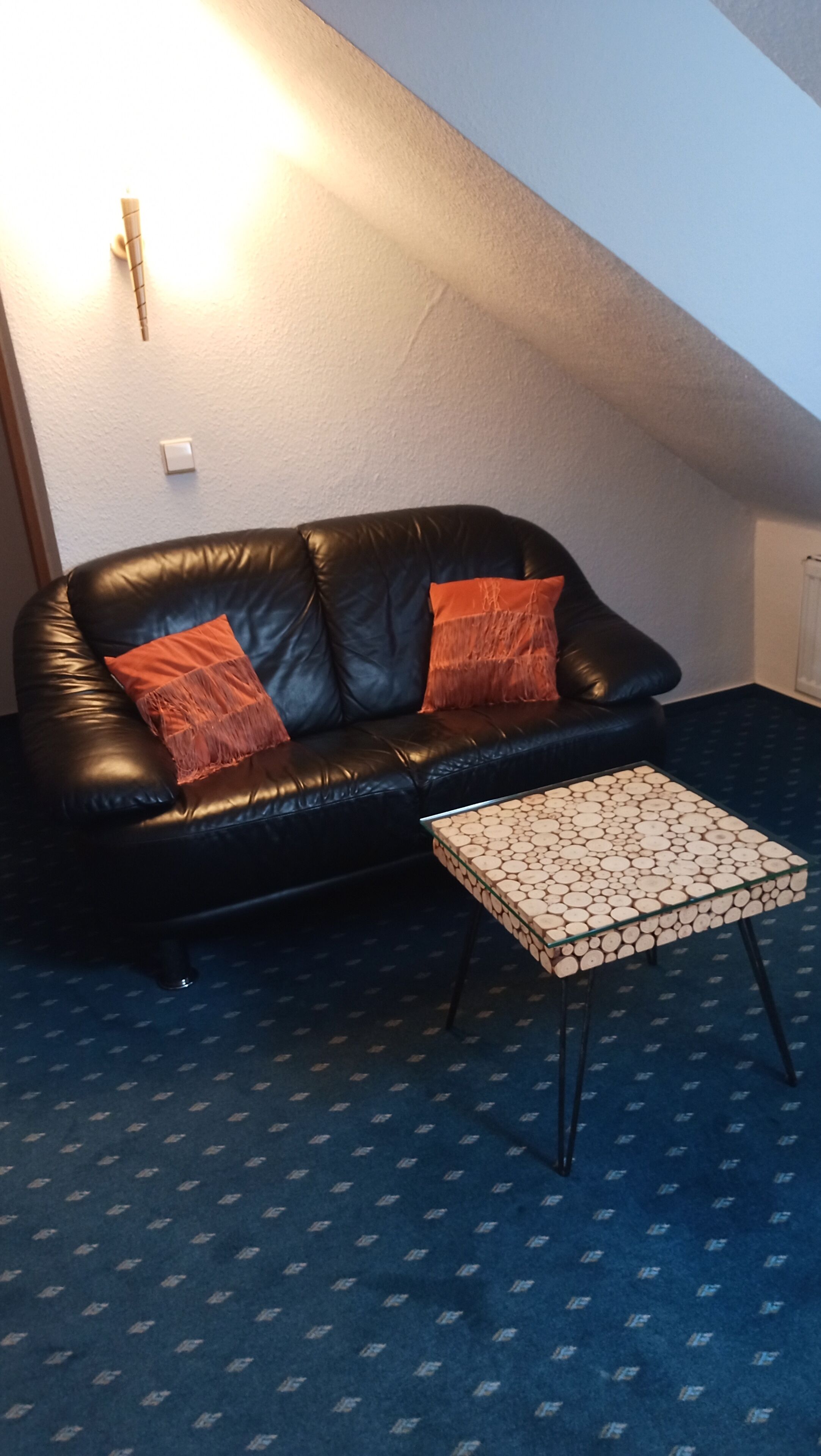Un sofá de cuero negro con dos almohadas anaranjadas, una mesa de centro de vidrio con diseño de madera y una alfombra estampada.