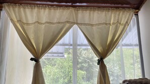 A room with a window covered by curtains tied with bows, a wooden table with a small item on it, and a view of greenery outside. - Butterfly and Bird Julio Tours-Ecolodge (Nanegal)