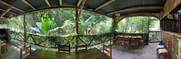 Un área de comedor al aire libre con estructuras de bambú, mobiliario de madera y abundante vegetación.