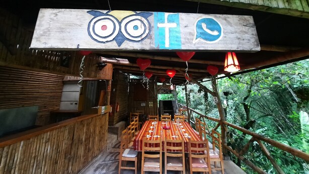 A rustic wooden structure with a table and chairs set up for dining, a hanging sign with owl and cross symbols, and a view of lush greenery outside. - Butterfly and Bird Julio Tours-Ecolodge (Nanegal)