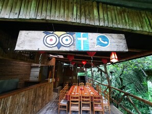 A rustic wooden structure with a table and chairs set up for dining, a hanging sign with owl and cross symbols, and a view of lush greenery outside. - Butterfly and Bird Julio Tours-Ecolodge (Nanegal)