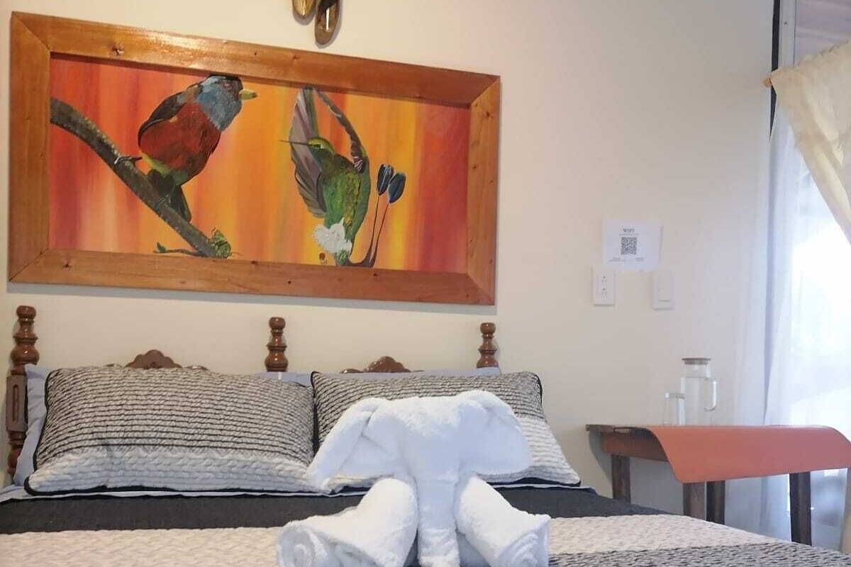 Een hotelkamer met een bed, kussens, een schilderij, een aan de muur bevestigd lampje en een raam met gordijnen.