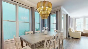 Un comedor con una mesa de madera, sillas blancas, una araña y ventanas amplias.