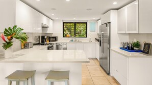 A modern kitchen with white cabinetry, a central island, stainless steel appliances, and a window with a view of greenery. - Topical ocean front 2-bedroom condo with AC in charming Koloa (Koloa)