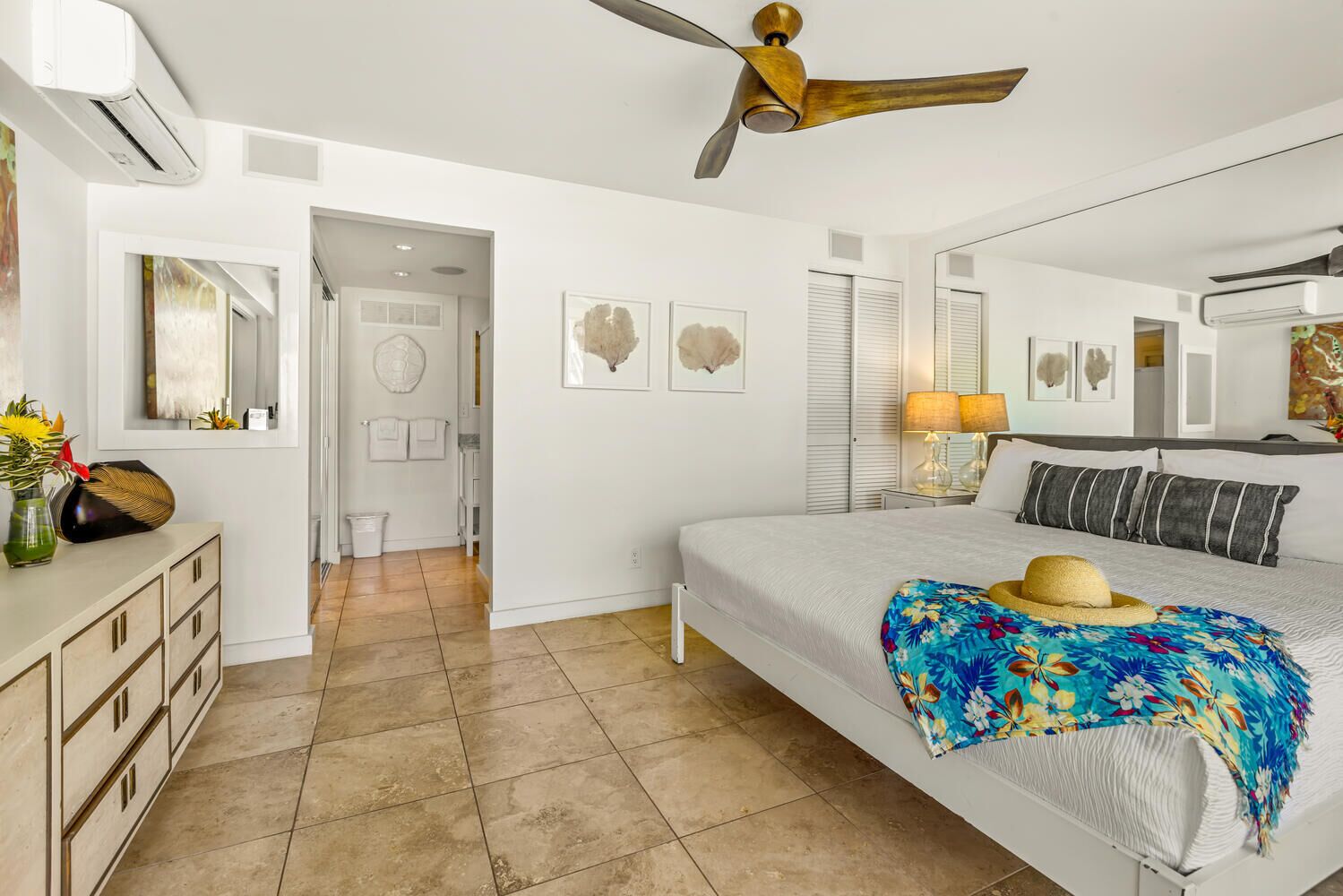 Quarto com cama, ventilador de teto, cômoda, espelho e porta que dá para outro espaço.