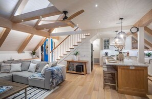 A modern loft with a high ceiling, exposed beams, a living area with a sofa and coffee table, a kitchen with a central island, and a staircase. - Neptune Ocean View Masterpiece (Encinitas)