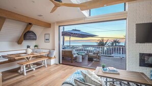 A modern interior with a wooden dining table, bench seating, and a large sliding glass door leading to a balcony with outdoor furniture. - Neptune Ocean View Masterpiece (Encinitas)