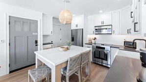 A modern kitchen with white cabinetry, a central island with seating, a hanging light fixture, and stainless steel appliances. - Luxury S Oceanfront Unit with Rooftop & Hot Tub + Sauna (San Diego)
