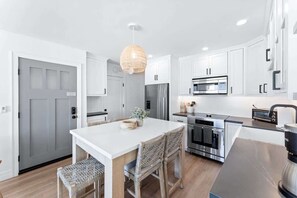 A modern kitchen with white cabinetry, a central island with seating, a hanging light fixture, and stainless steel appliances. - Luxury S Oceanfront Unit with Rooftop & Hot Tub + Sauna (San Diego)