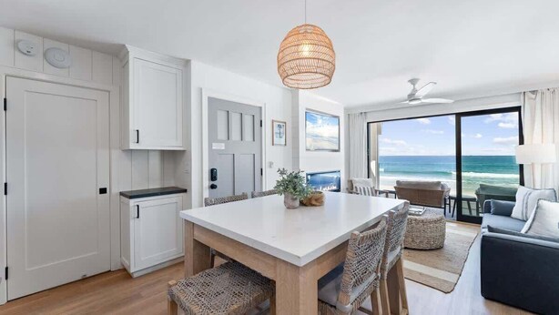 A modern kitchen with white cabinetry, a dining area with wicker chairs, and a view of the ocean through large windows. - Luxury oceanfront fourplex with roof deck, sleeps 22 (San Diego)