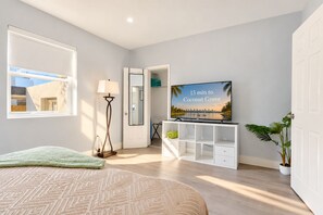 Un dormitori amb un llit, un televisor sobre un suport, una làmpada de peu, una porta que dona a un bany i una finestra amb vista a un edifici.