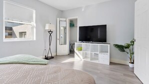 Une chambre à coucher moderne avec un lit, un téléviseur posé sur un meuble blanc, une lampe sur pied et une plante en pot.
