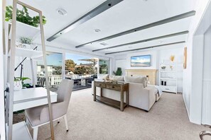 A modern living room with a white sofa, a coffee table, and a TV. There is a balcony with outdoor furniture and plants. - Stunning BayVIEW Home with Epic Waterfront Views! (Newport Beach)