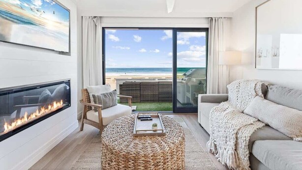 A living room with a fireplace, a sofa, a round woven ottoman, and a view of the beach through large glass doors. - Luxury Ground N Oceanfront Unit with Patio Lounge (San Diego)