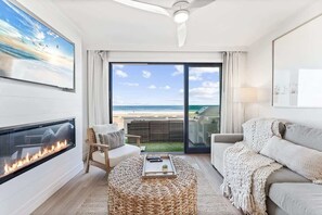 Ein Wohnzimmer mit Kamin, einer Couch, einem runden gewebten Hocker und Blick auf den Strand durch große Glasüren.