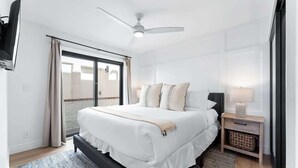 A bedroom with a bed, a ceiling fan, a TV, a nightstand, and a view through a window. - Luxury Ground N Oceanfront Unit with Patio Lounge (San Diego)