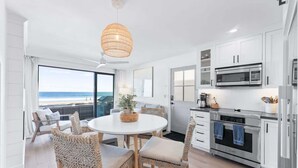 A modern kitchen with white cabinets, stainless steel appliances, and a dining area with wicker chairs and a round table. - Luxury Ground N Oceanfront Unit with Patio Lounge (San Diego)