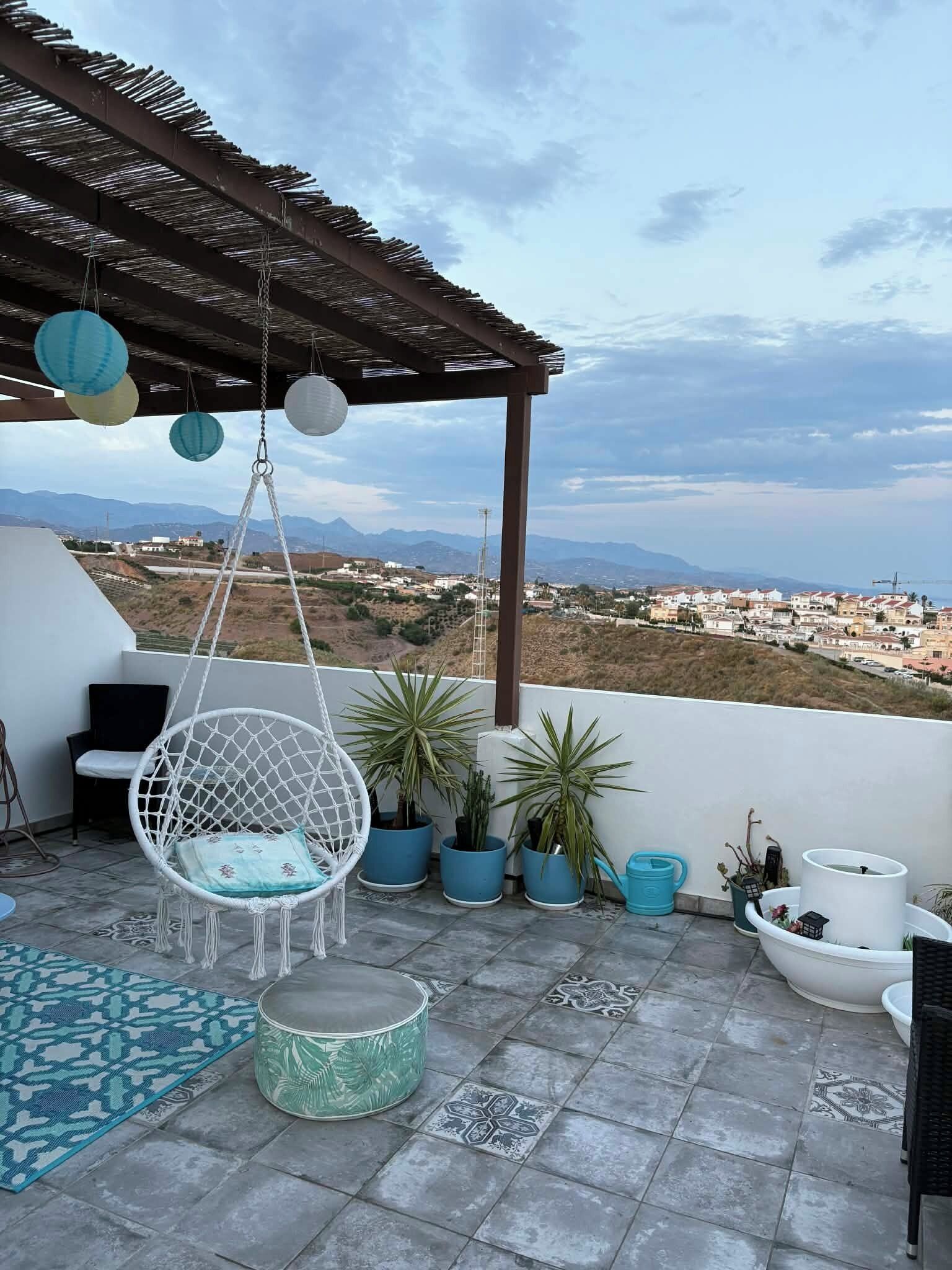 Un patio con una silla colgante, plantas en macetas, un tapete estampado y vistas a montañas y edificios a lo lejos.