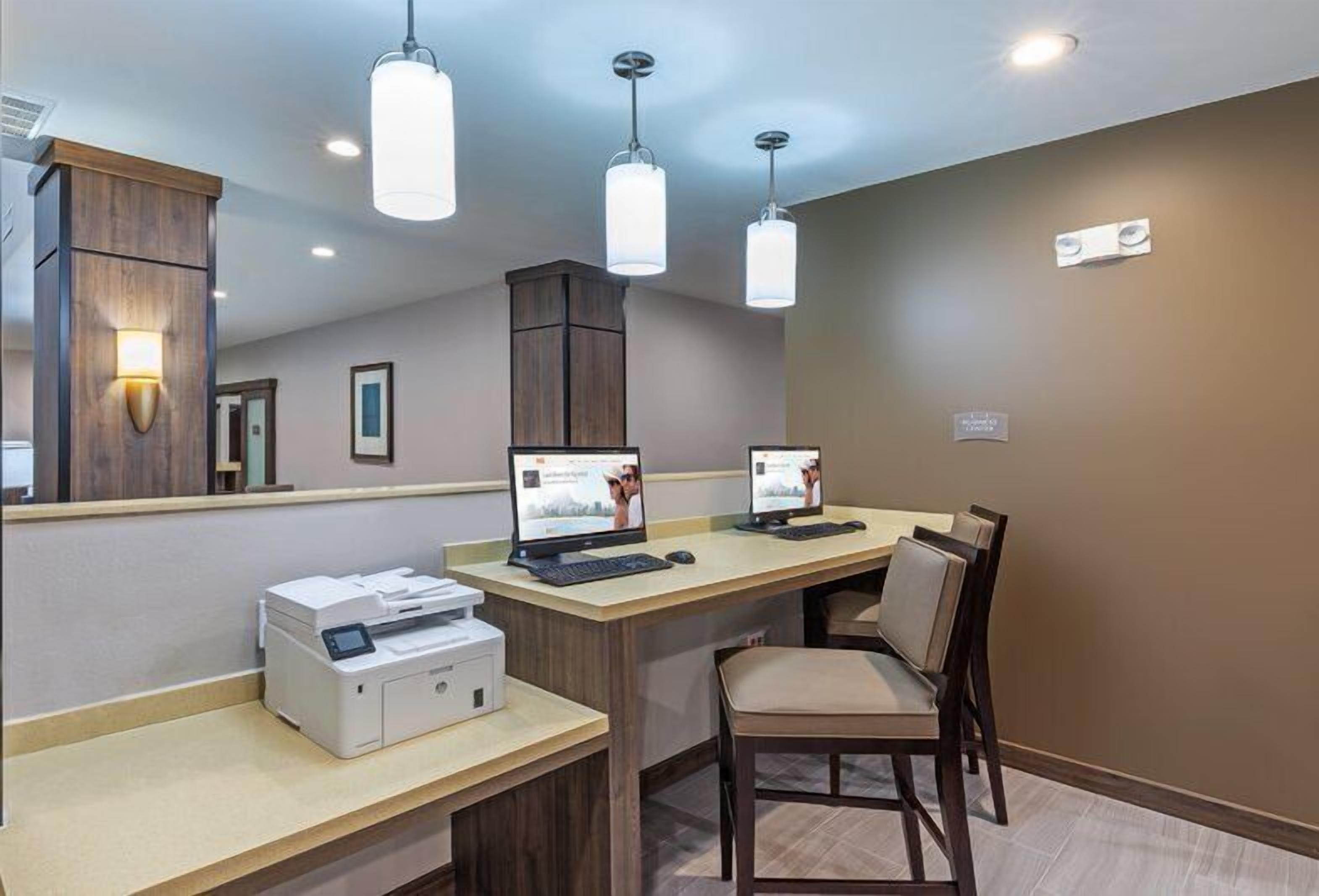 Habitación de hotel con escritorio, dos computadoras, una impresora, una silla e iluminación colgante.