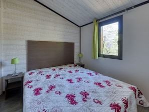 A bedroom with a bed, a nightstand, a lamp, a window with curtains, and a wooden ceiling.