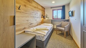 A bedroom with a wooden headboard, a bed, a desk, a chair, a window with a view, and a TV on the wall. - Hotel Bouda Jana (Pec pod Sněžkou)