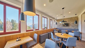 A modern dining area with wooden tables and chairs, large windows, and a stone wall. - Hotel Bouda Jana (Pec pod Sněžkou)
