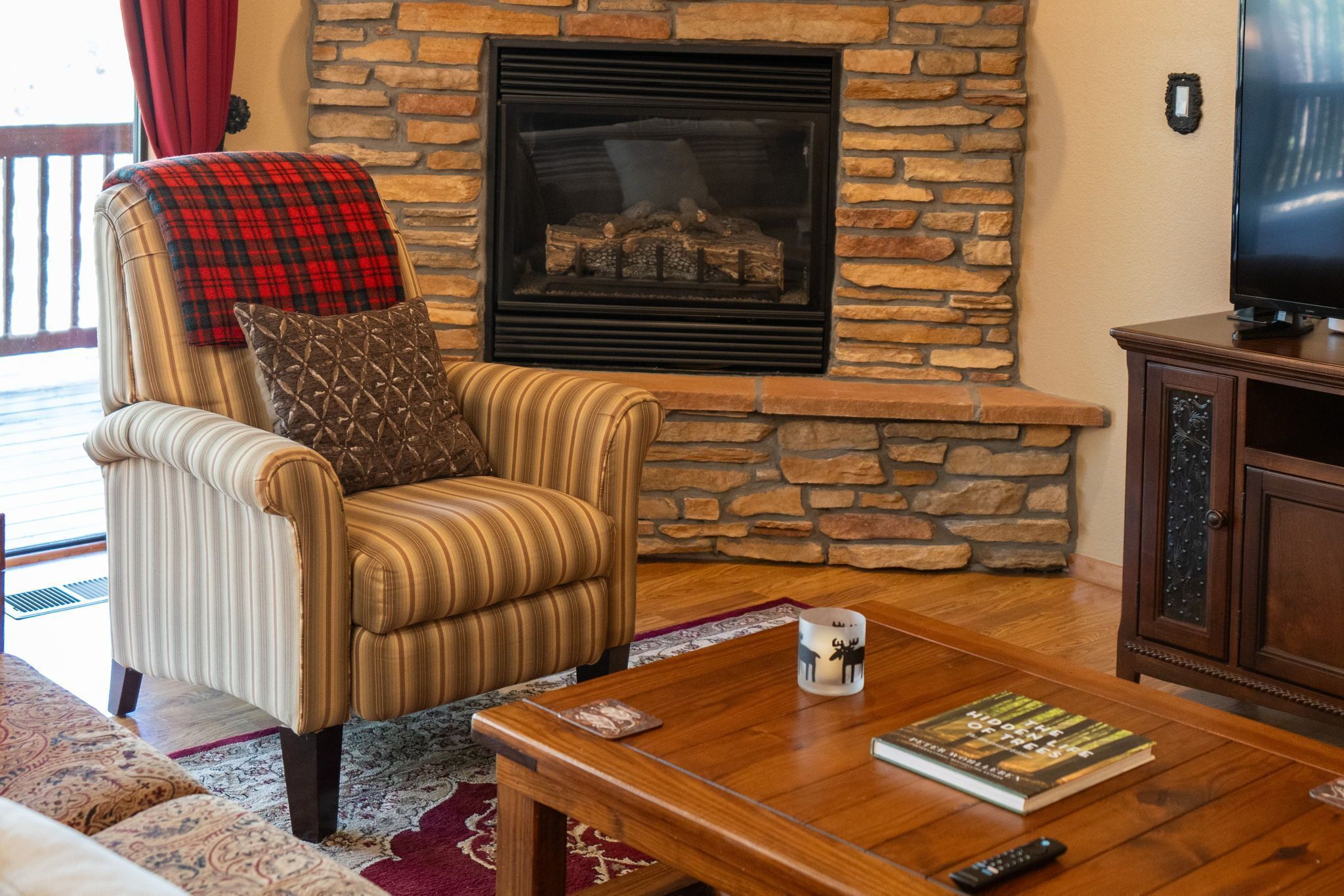 A living room with a stone fireplace, a striped armchair, a wooden coffee table, and a television.