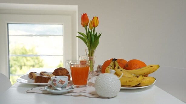 Een ontbijttafel met gebak, een glas sinaasappelsap, een vaas met tulpen en een bord met bananen en mandarijnen.