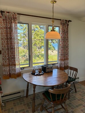 A dining area with a round wooden table, two chairs, a hanging light fixture, and a view of trees and water through large windows. - Manset oceanfront 3-bedroom home with sweeping views (Southwest Harbor)