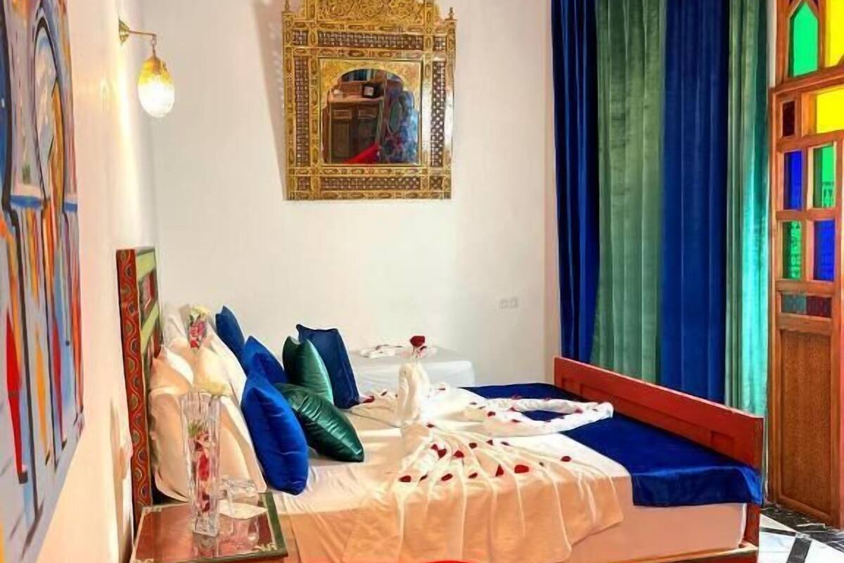 Ein Zimmer mit einem Bett, einem roten Sessel, einer Kristallleuchtre, einem Spiegel und einem farbenfrohen Fenster.
