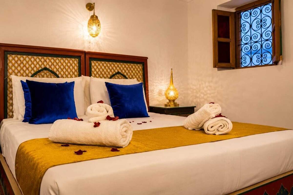 Ein Hotelzimmer mit Bett, Kissen, Nachttisch, Lampe und einem Fenster mit schmiedeeisernem Zierrat.
