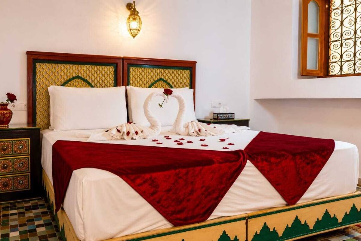 Ein Bett mit roter Bettdecke, ein dekoratives Kopfteil, ein Nachttisch mit Schubladen, eine Vase mit einer roten Rose und eine wandmontierte Leuchte.