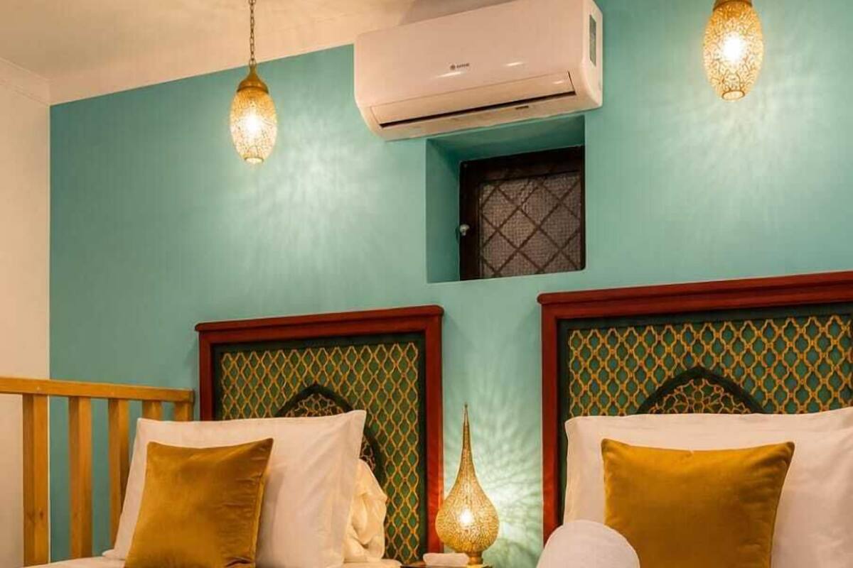 Ein Hotelzimmer mit zwei Betten, einer fest installierten Klimaanlage, einer dekorativen Lampe und einem kleinen Fenster mit Gittermuster.