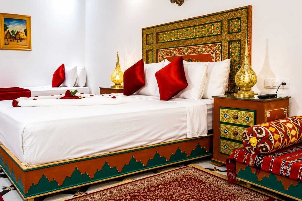 A hotel room with a large bed, two bedside tables, a nightstand, a decorative headboard, a wall-mounted painting, and a patterned rug.