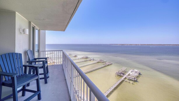 A balcony with two Adirondack chairs, a view of the ocean, and a pier with boats. - Bayfront Bliss in Gated Tristan Towers – 2 Bed, 2 Bath, Sleeps 4 (Pensacola Beach)