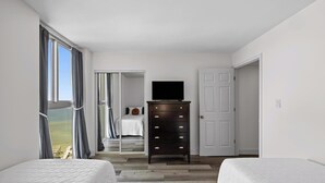 A bedroom with two beds, a dresser, a TV, and a view of a green field through a window. - Bayfront Bliss in Gated Tristan Towers – 2 Bed, 2 Bath, Sleeps 4 (Pensacola Beach)