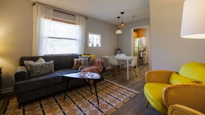 A living room with a sofa, a small table, and a yellow chair. There is a dining area with a table and chairs, and a kitchen in the background.