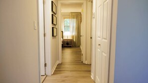A narrow hallway with wooden flooring, white walls, and a door at the end leading to a room with a window and curtains.