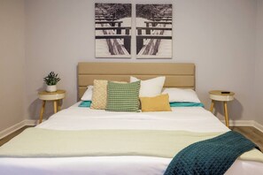 A bed with a beige headboard, multiple pillows, a white bedspread, and a teal throw blanket. There are two bedside tables with small potted plants.