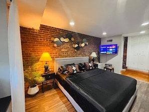 A bedroom with a brick wall, a bed with a black bedspread, a TV on the wall, and a plant on the nightstand. - Ground floor flat at Virginia Highlands (Atlanta)