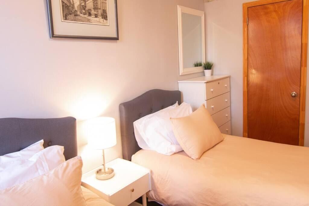 A bedroom with two beds, a bedside table with a lamp, a dresser, a mirror, and a door.